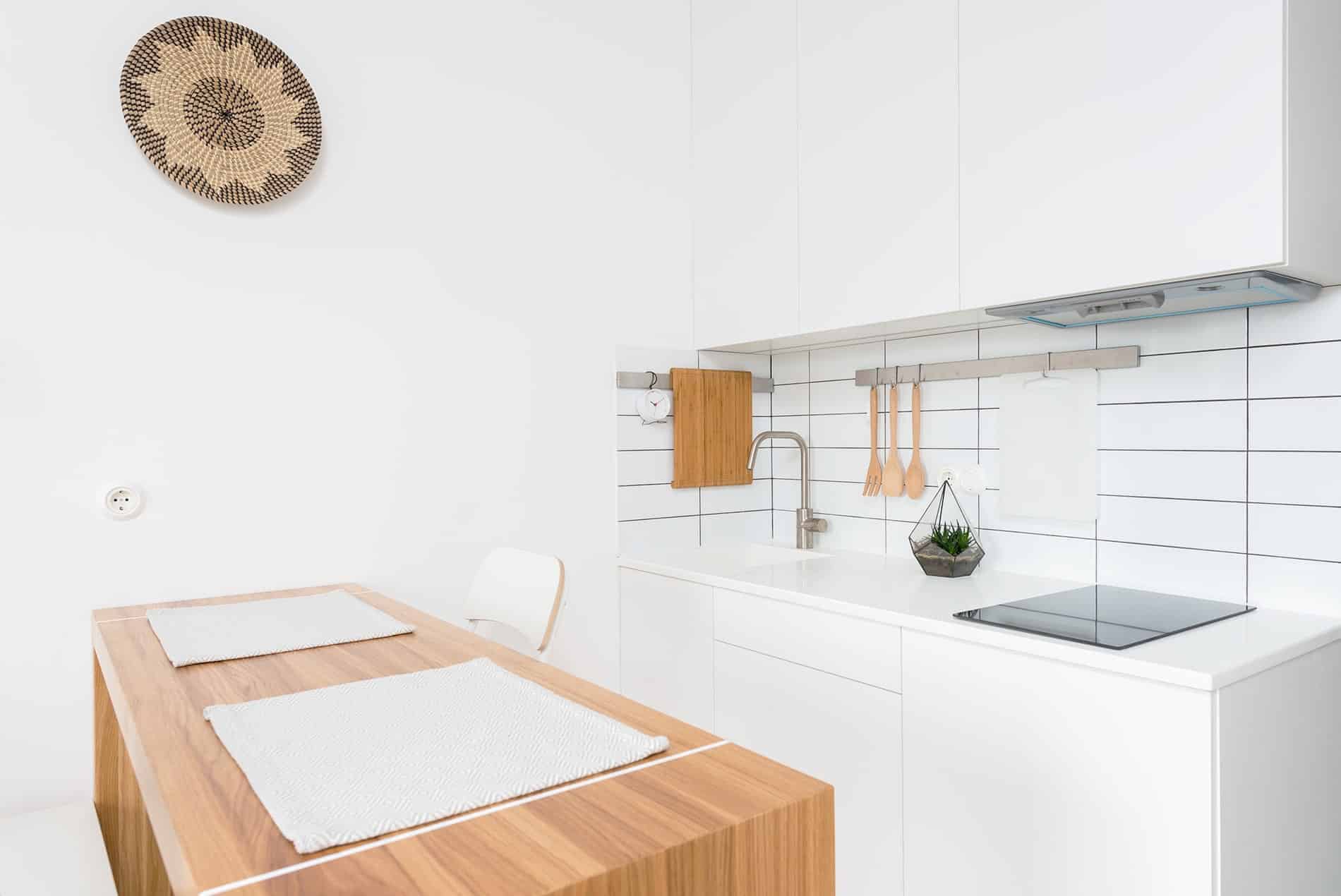Minimalist kitchen with white cabinets, wooden countertop, and decorative wall basket. Simple design featuring tiled backsplash, plants, and cooking utensils on display.